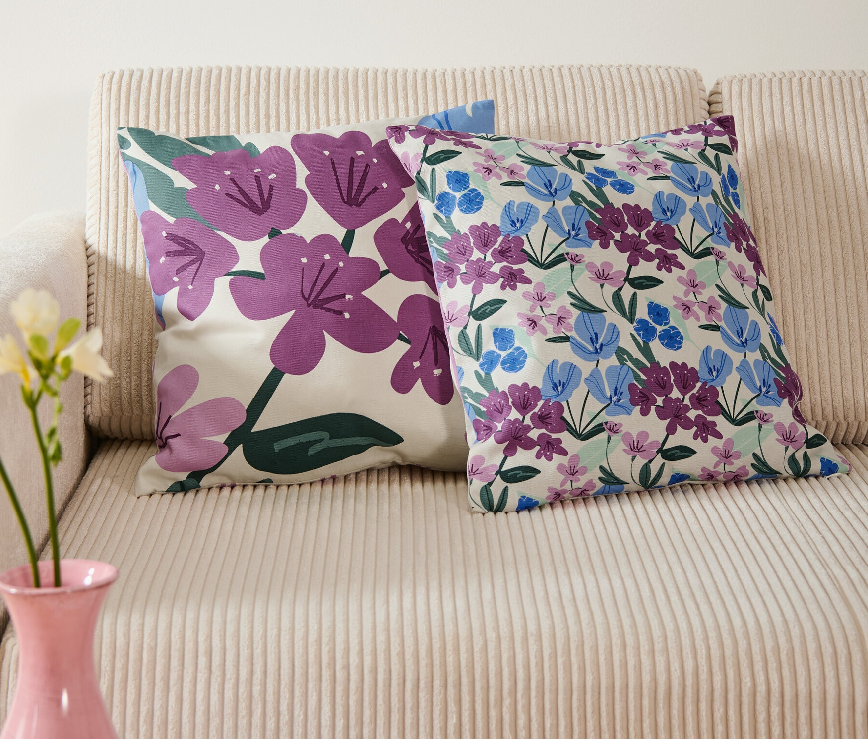 Zwei Dekokissenbezüge mit Blumenmuster liegen auf einem beigen Sofa, darüber hängt ein blaues Bild mit Blumen in einer Vase.