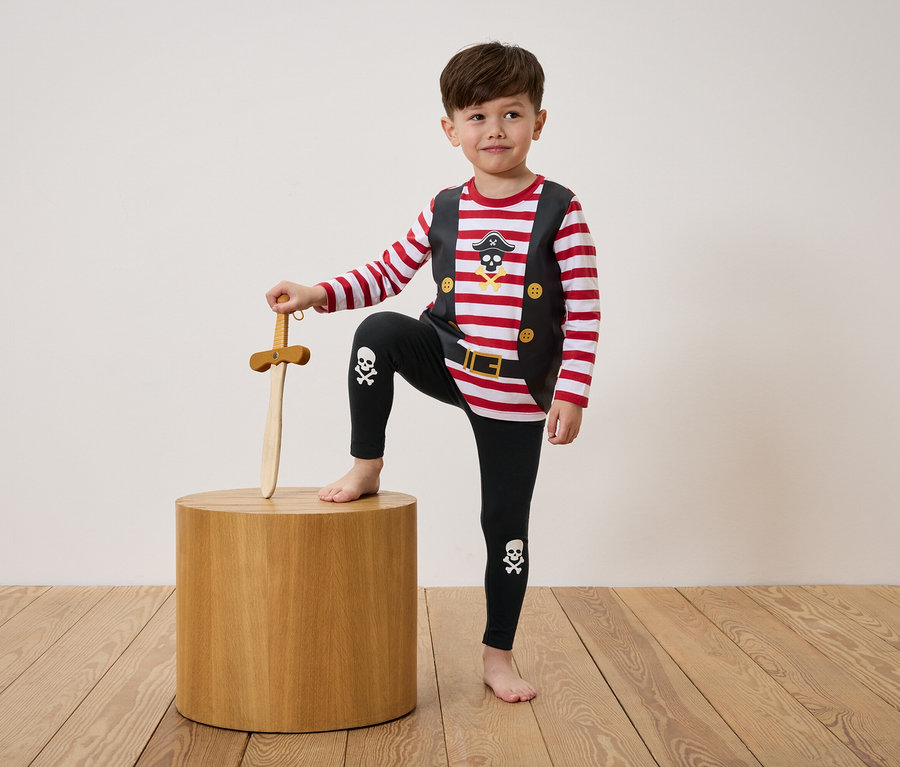 Junge im Piratenkostüm steht mit einem Bein auf einem Holzhocker und hält ein Holzschwert.