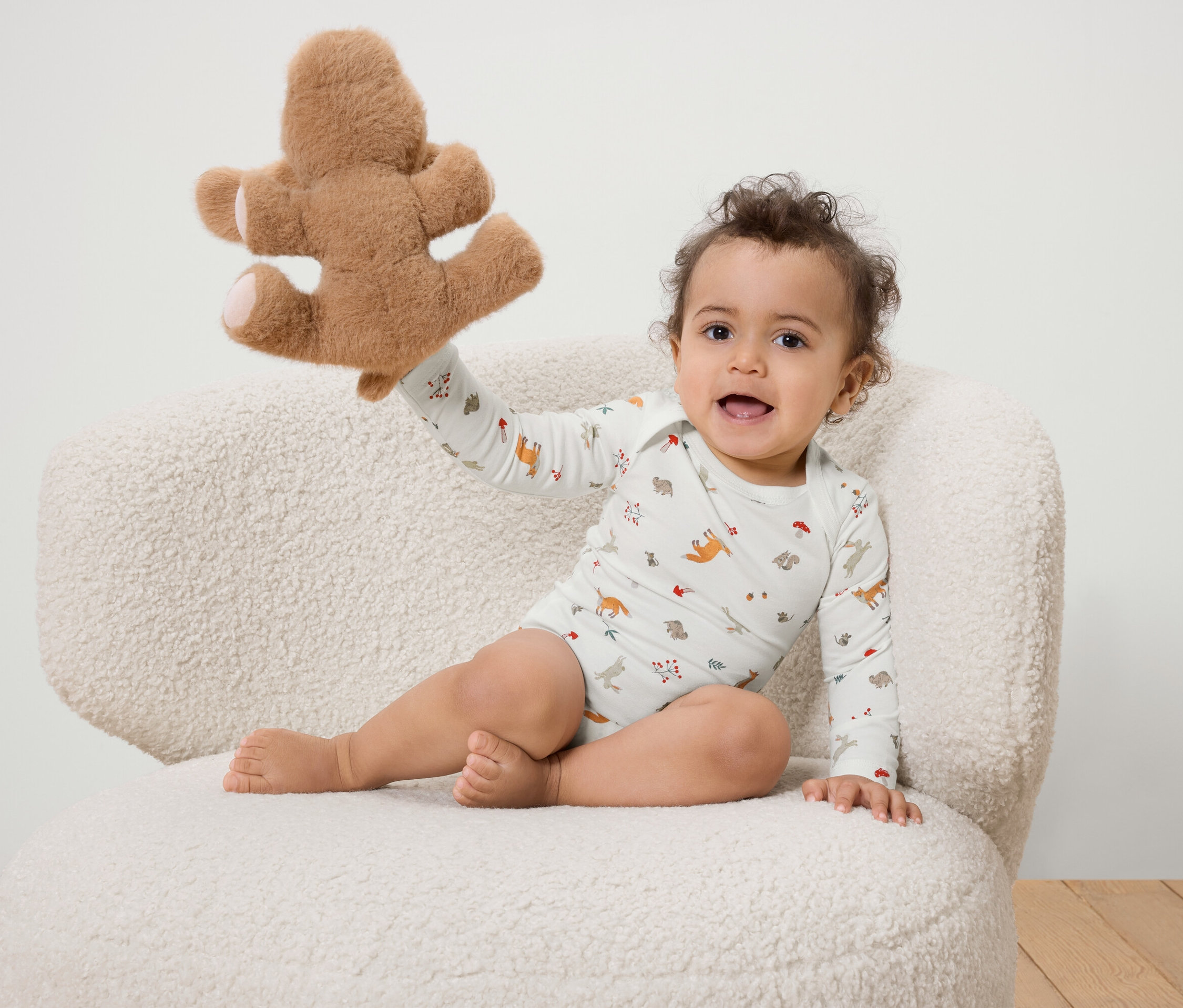 Ein lächelndes Baby sitzt auf einem weißen, flauschigen Stuhl und hält einen Teddybären.