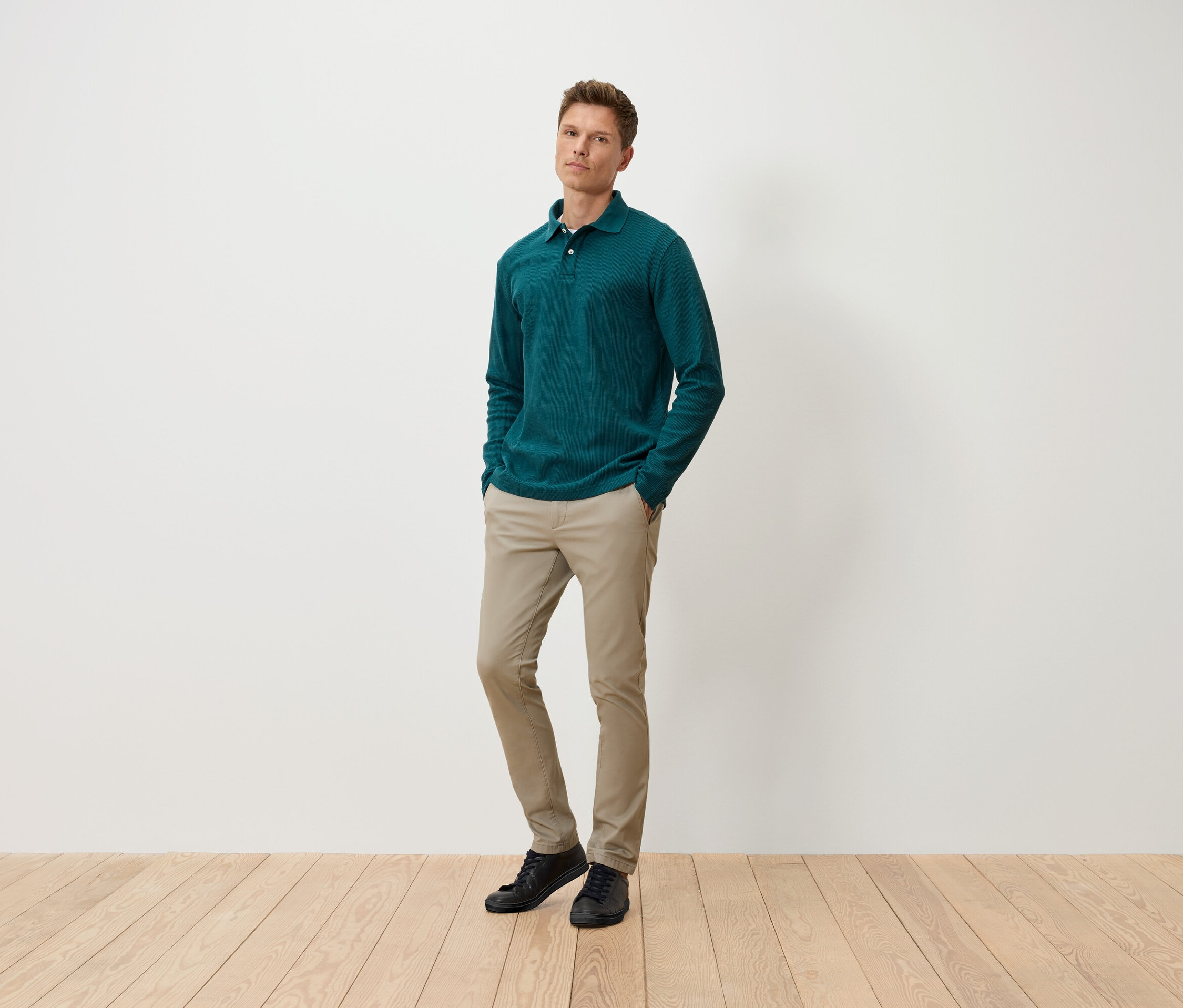 Ein Mann steht mit den Händen in den Hosentaschen und trägt ein grünes Langarm-Poloshirt, beige Hosen und schwarze Schuhe.