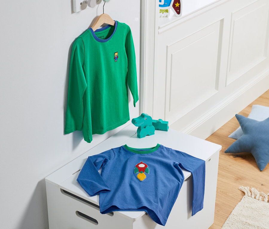 Ein grünes Langarmshirt aus einem 2er-Set hängt auf einem Bügel, ein blaues Langarmshirt liegt auf einer weißen Truhe.