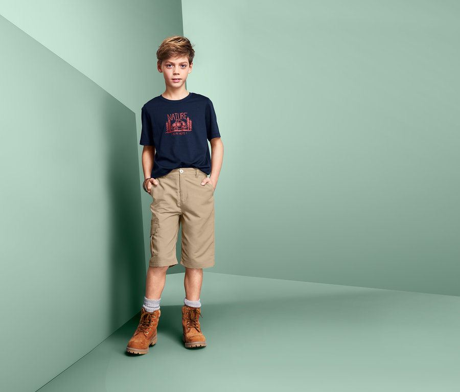Ein Junge posiert in beigen 2-in-1-Funktionsshorts, marineblauem T-Shirt und braunen Stiefeln.