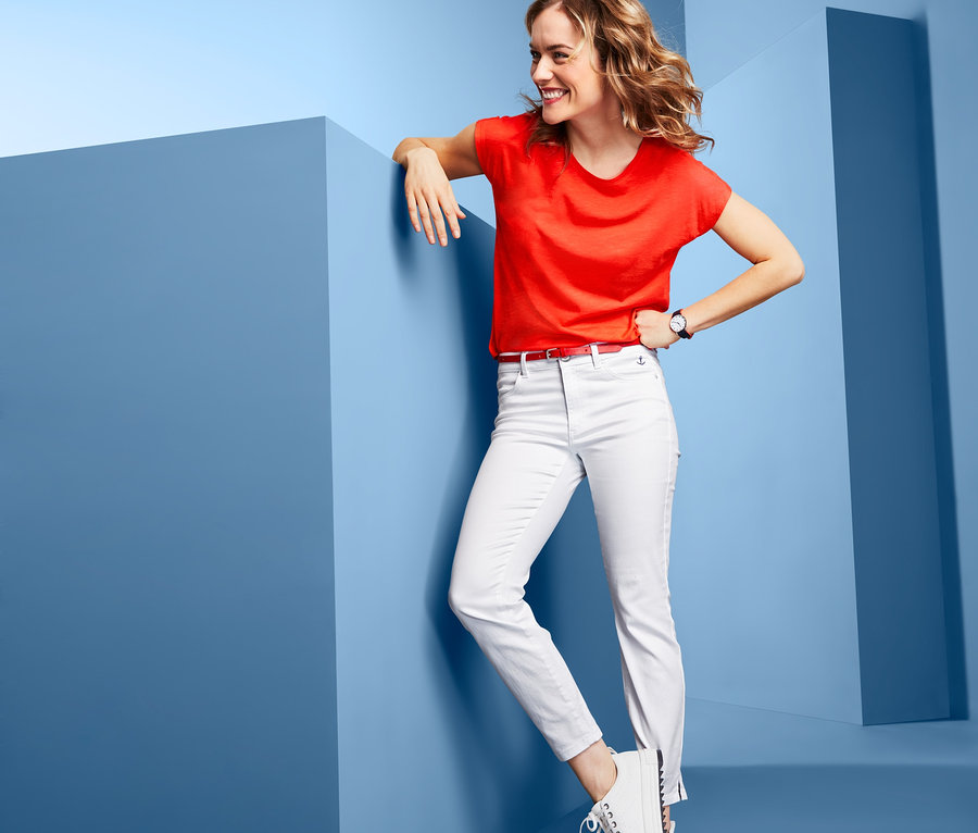 Lächelnde Frau lehnt an einem blauen Block, trägt ein rotes T-Shirt und eine knöchellange Hose.