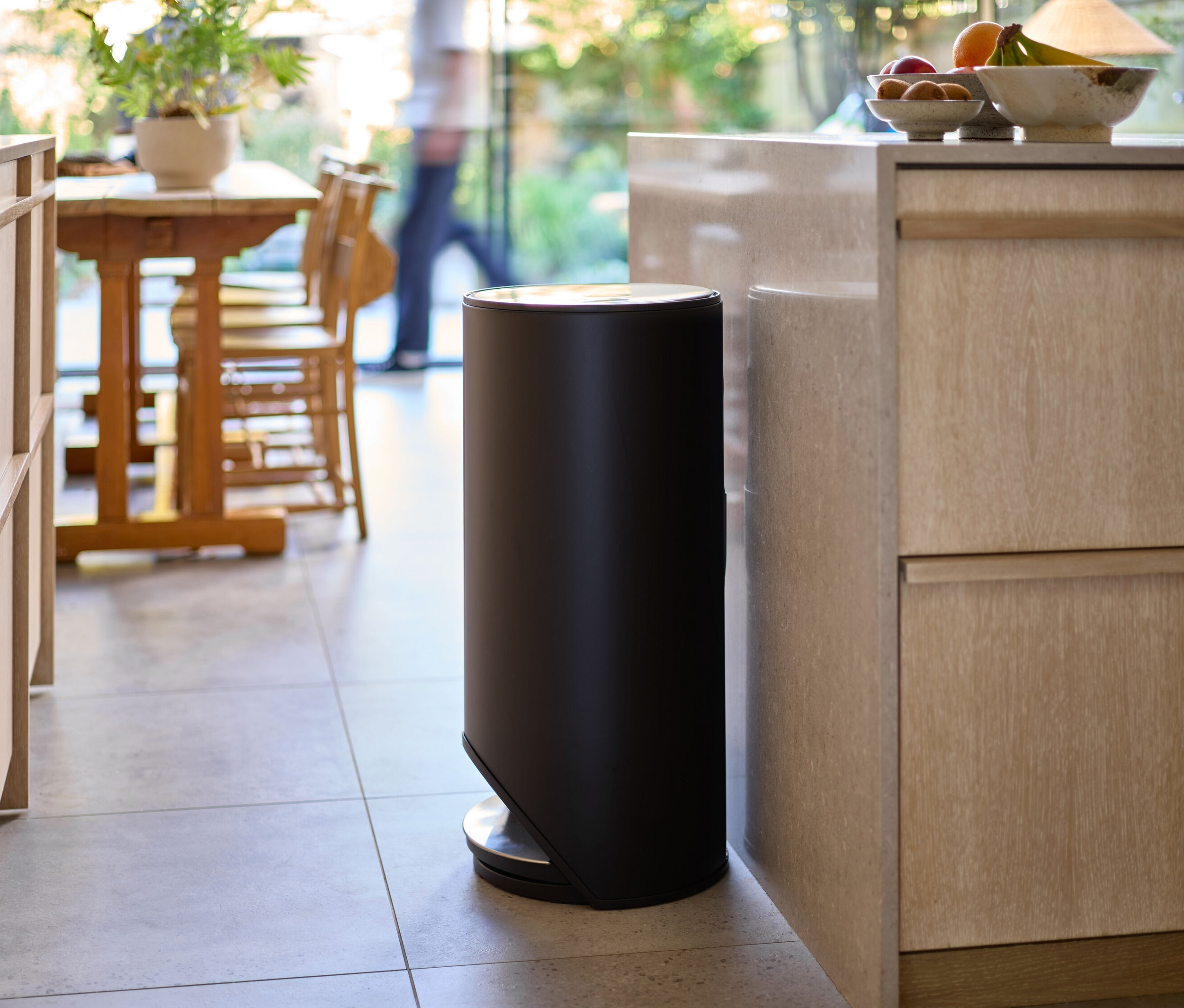 Ein schwarzer Abfalleimer steht in einer Küche neben einem Schrank. Im Hintergrund sind ein Tisch mit Stühlen und eine Person zu sehen.