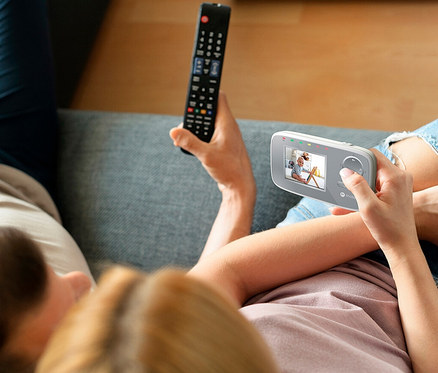 Ein Mann und eine Frau sitzen auf einer Couch. Die Frau hält eine Fernbedienung und ein Video-Babyphone "VM482" in der Hand.