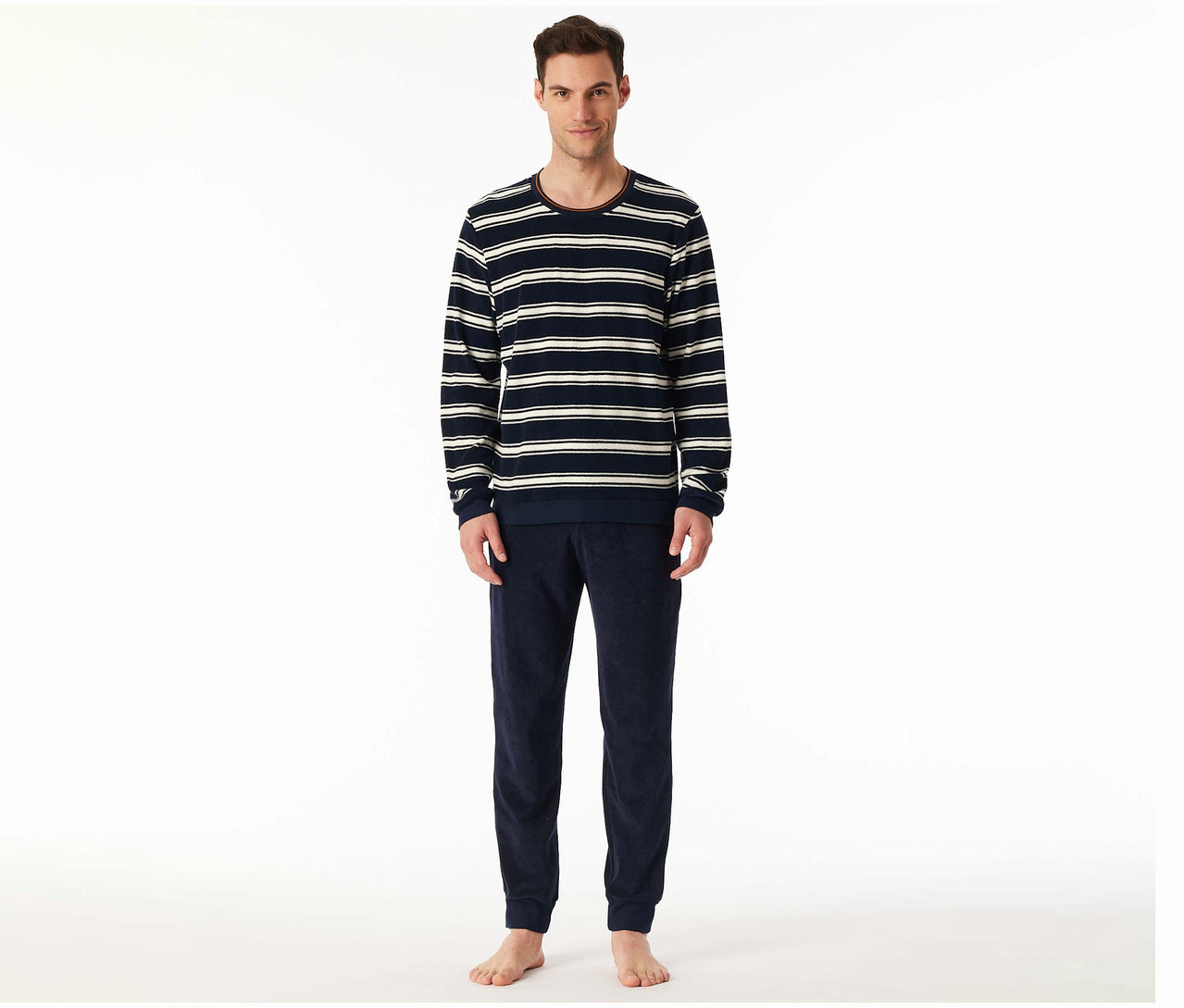 Ein lächelnder Mann steht in einem SCHIESSER Frottee-Pyjama-Set. Das langärmelige Oberteil ist marineblau mit weißen Streifen und rotem Rand, dazu eine unifarbene marineblaue Hose.