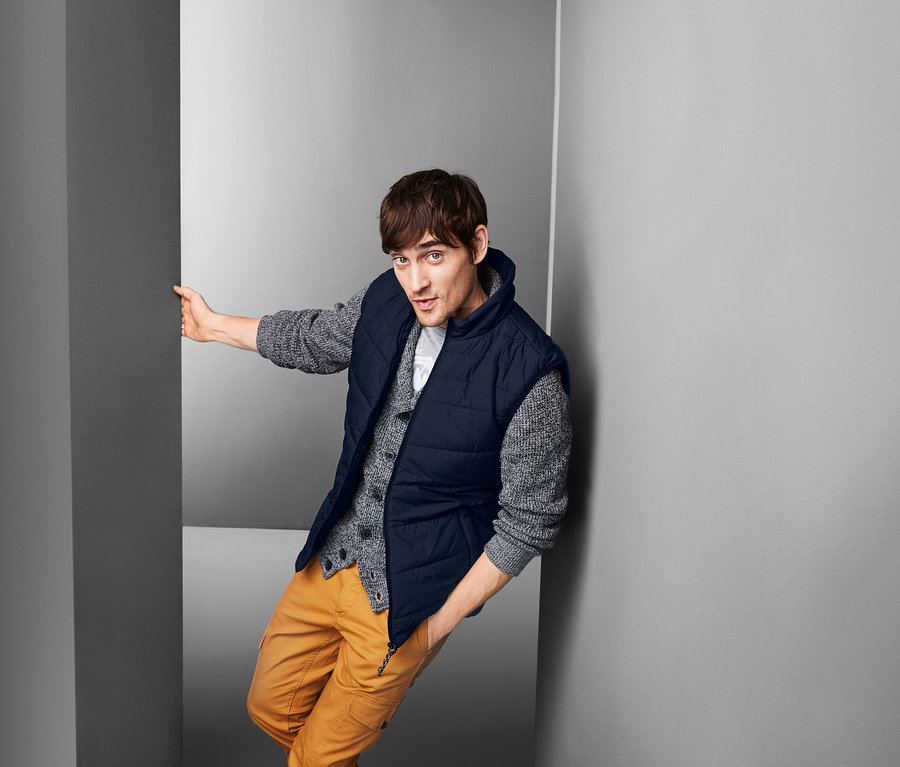 Mann lehnt an Wand und trägt Steppweste, Pullover und Hose.