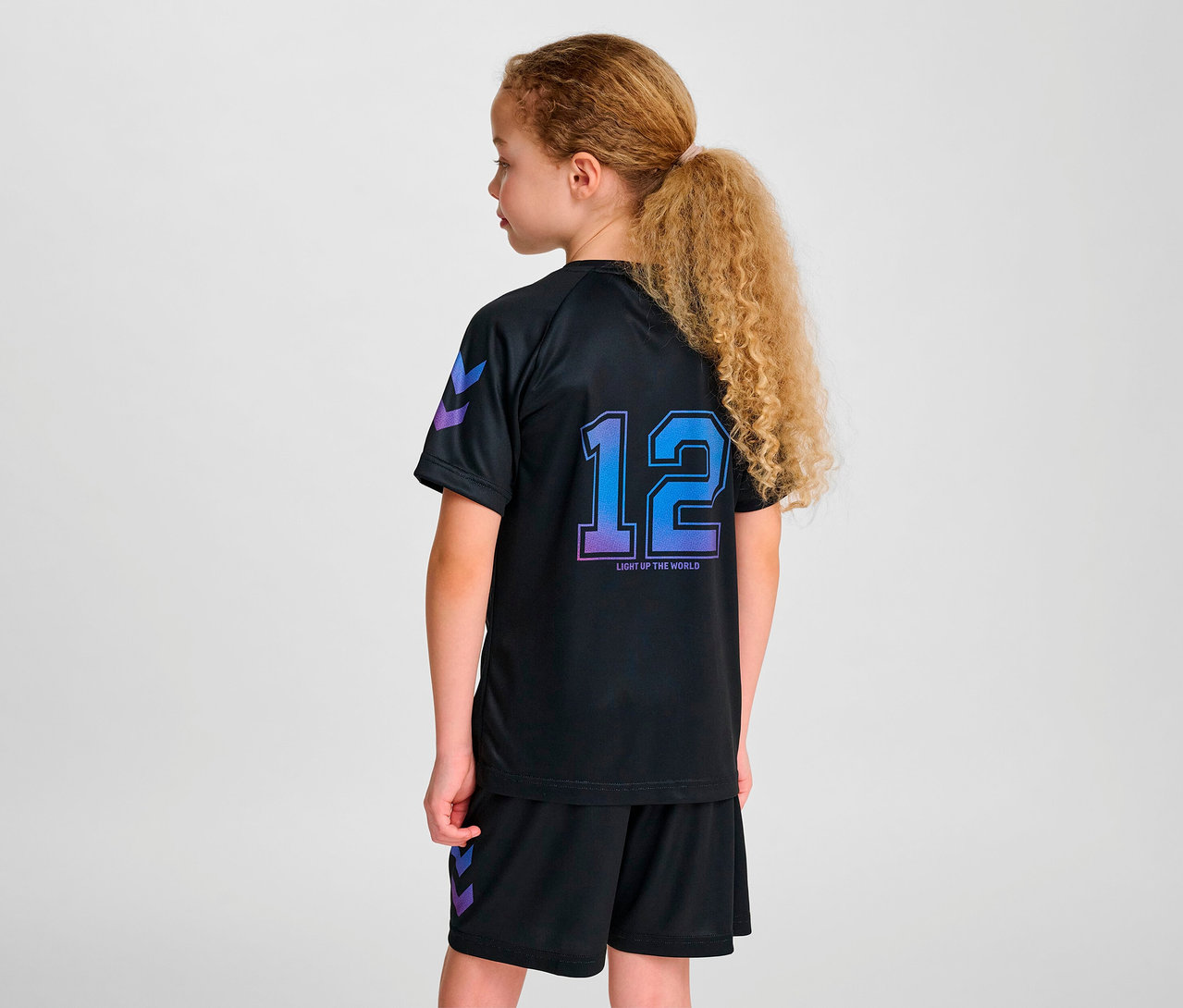 Junges Mädchen trägt ein schwarzes Sportset mit der Nummer 12 auf der Rückseite des HUMMEL HMLJR Shimmer T-Shirts S/S.