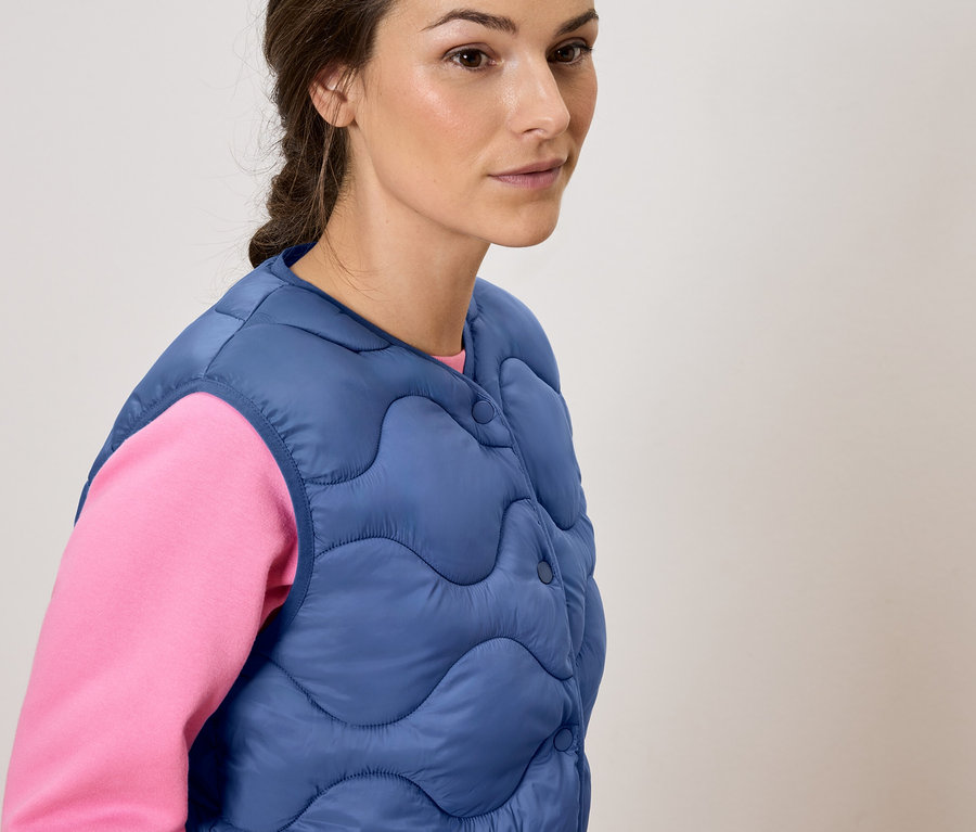 Frau trägt eine blaue Steppweste über einem rosa Oberteil.