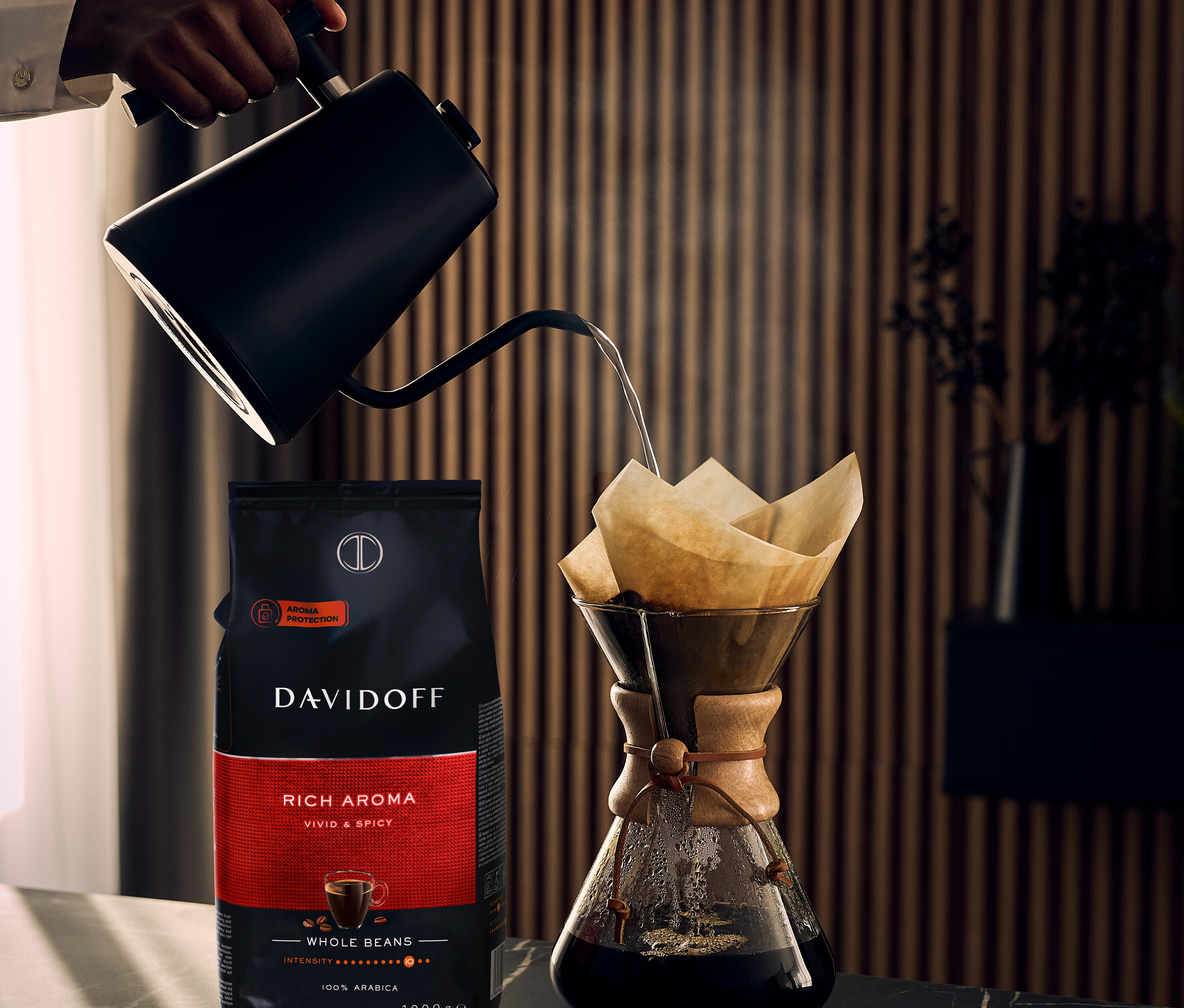 Hand gießt Wasser aus einem schwarzen Kessel in einen Kaffeefilter neben einer Packung DAVIDOFF CAFÉ Rich Aroma.