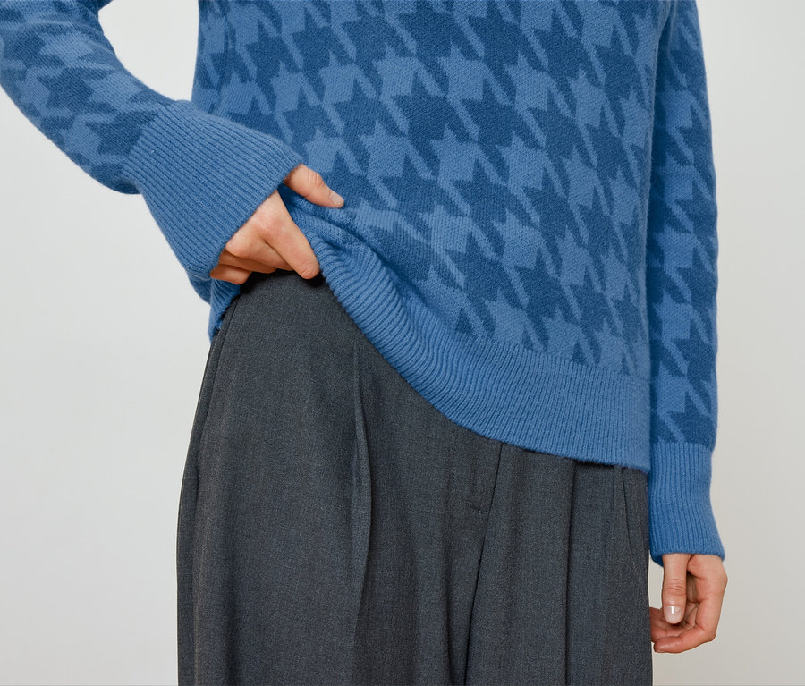 Person zupft an einem blauen Pullover mit Hahnentrittmuster und trägt eine graue Hose.