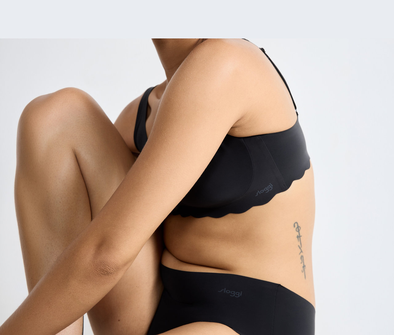 Eine Person trägt einen schwarzen sloggi ZERO Microfibre 2.0 Soft bra mit Wellenkante und passende schwarze sloggi Slips, sitzend mit angewinkelten Knien und verschränkten Armen. Ein Tattoo ist an der Seite sichtbar.