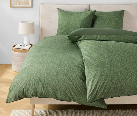 Grüne Bettwäsche mit Muster auf einem Bett mit beigem Kopfteil. Auf dem Nachttisch stehen eine Lampe und eine Brille.