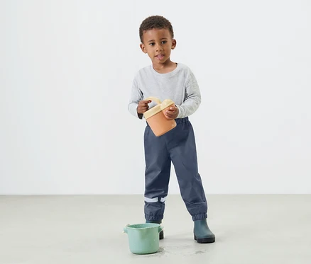 Ein kleiner Junge hält einen orangefarbenen Eimer in seinen Händen und trägt dabei blaue Regenhosen und Gummistiefel.