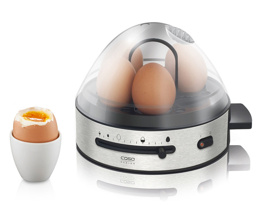 Vier braune Eier in einem Eierkocher aus Edelstahl mit transparentem Deckel und ein Ei in einem weißen Eierbecher.