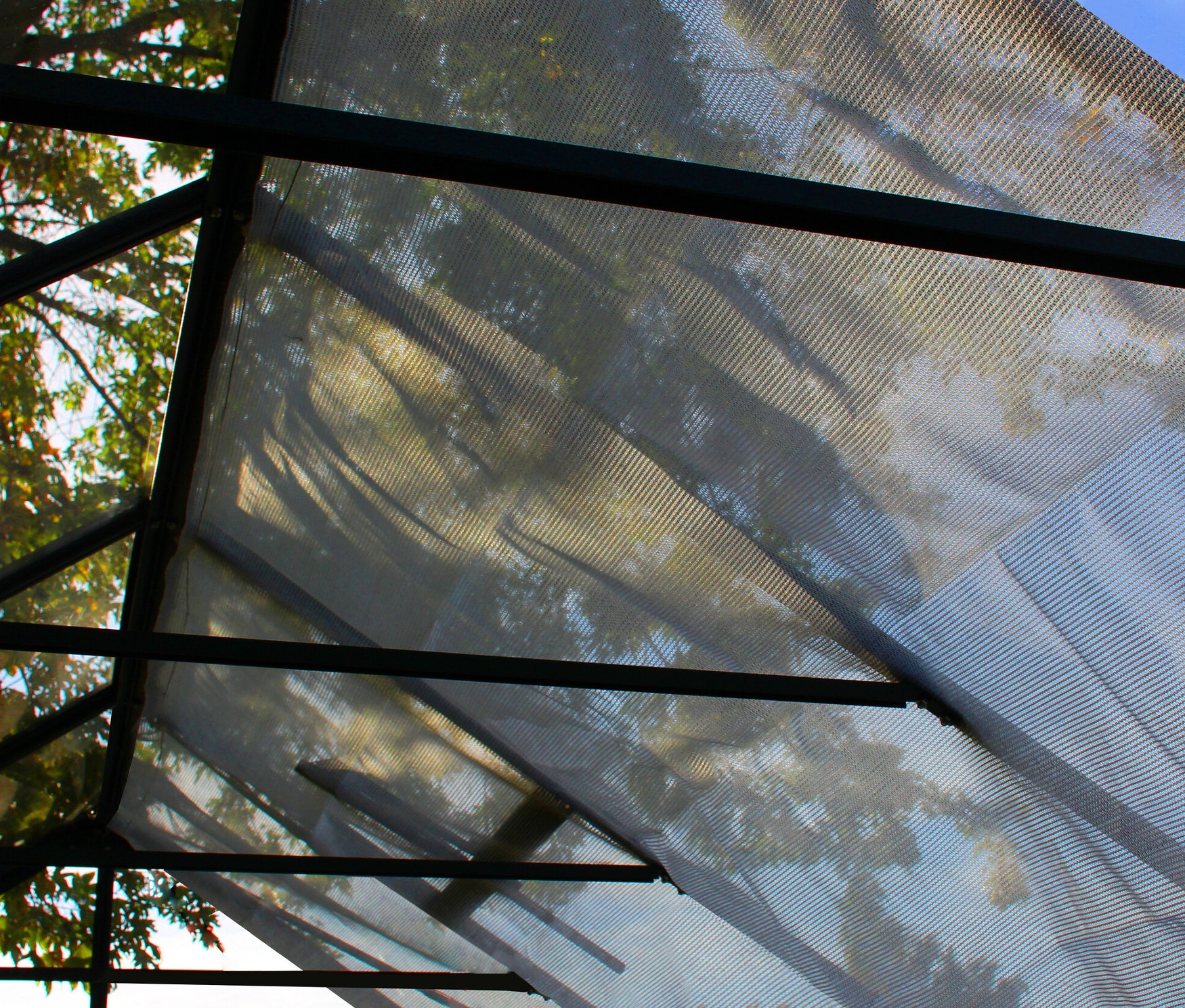 Blick von unten auf ein Vitavia Schattiergewebe, das an einer Metallstruktur befestigt ist, mit durchscheinenden Bäumen und Himmel.