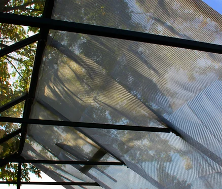 Blick von unten auf ein Vitavia Schattiergewebe, das an einer Metallstruktur befestigt ist, mit durchscheinenden Bäumen und Himmel.