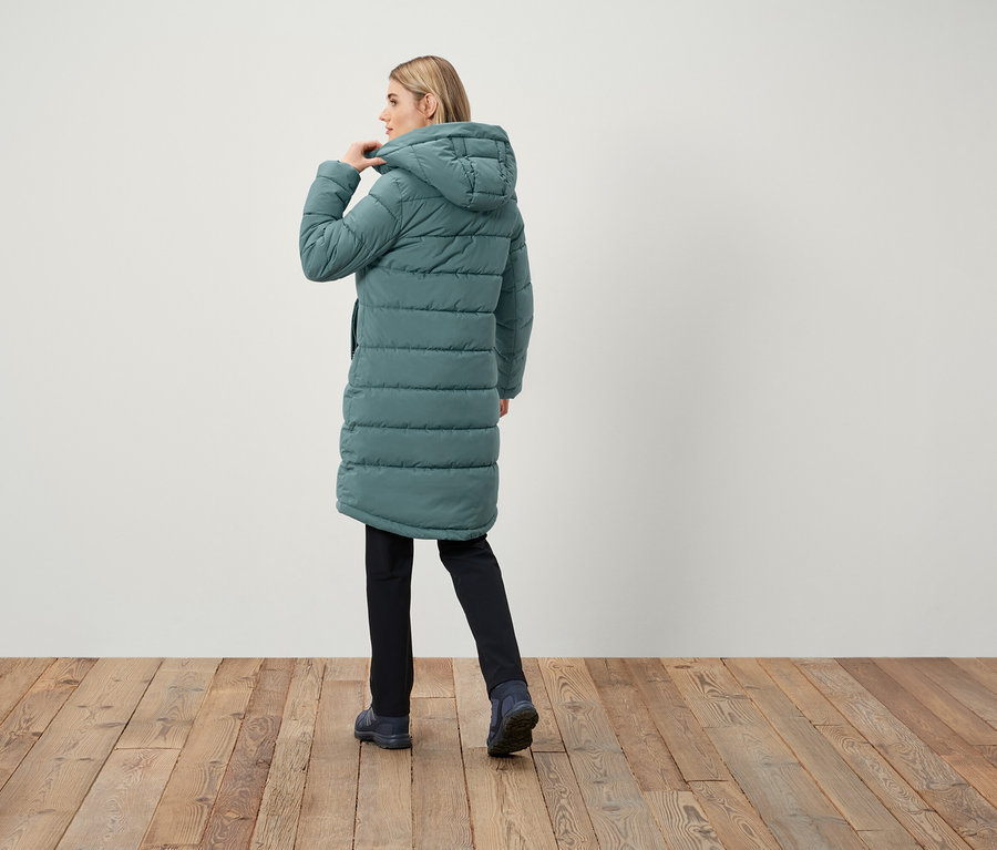 Frau trägt einen langen, gesteppten Mantel mit Kapuze, mit dem Rücken zur Kamera.