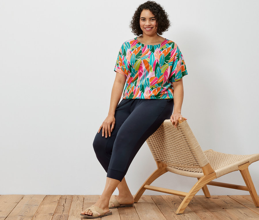 Eine Frau sitzt auf einem Korbstuhl und trägt eine dunkelblaue 3/4-Jerseyhose und ein überschnittenes Blusenshirt mit Alloverprint.