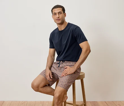 Mann sitzt auf einem Hocker und trägt ein marineblaues T-Shirt und gemusterte Shorts.