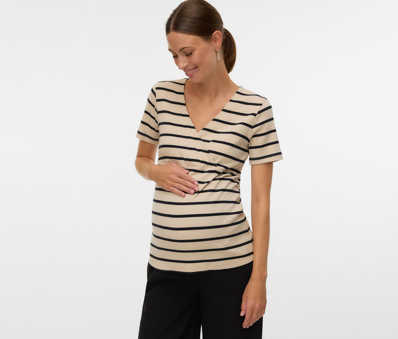 Frau mit Pferdeschwanz trägt ein gestreiftes MAMALICIOUS Umstands-T-Shirt »MLLANE TESS« und schwarze Hose, während sie ihren Bauch berührt.
