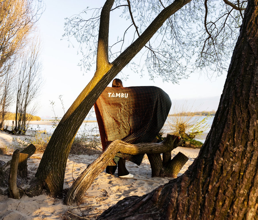 Person steht am Strand unter einem Baum, gehüllt in eine Tambu Campingdecke »KAMBALA« mit Punktmuster und großem Logo.