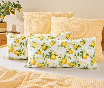 Auf einem Bett liegen zwei Kissen mit Zitronenmuster vor zwei gelben Kissen. Links steht eine Zitronenpflanze neben einer Kerze.