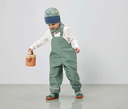 Ein Kind in grüner Latzhose, Mütze und Halstuch mit Dinosaurier-Muster hält eine Gießkanne in der Hand.