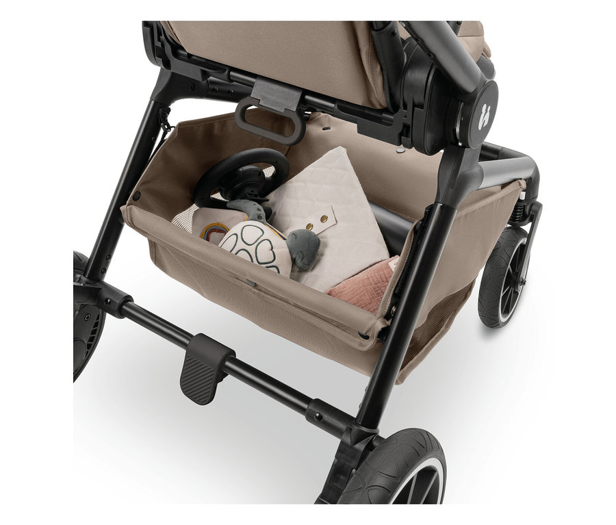 In der Ablage eines Kinderwagens liegen ein Lenkrad, ein Spielzeugschildkröte, eine beigefarbene Decke und ein rosa Musselintuch.