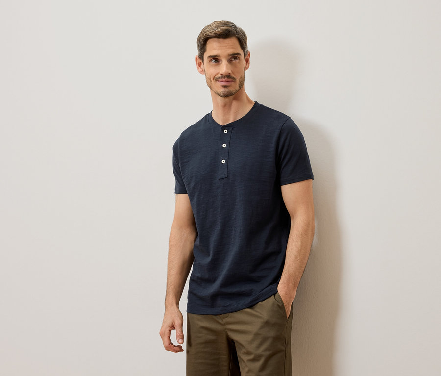 Mann mit blauem Henley-Shirt und khakifarbenen Hosen, der an einer weißen Wand posiert.