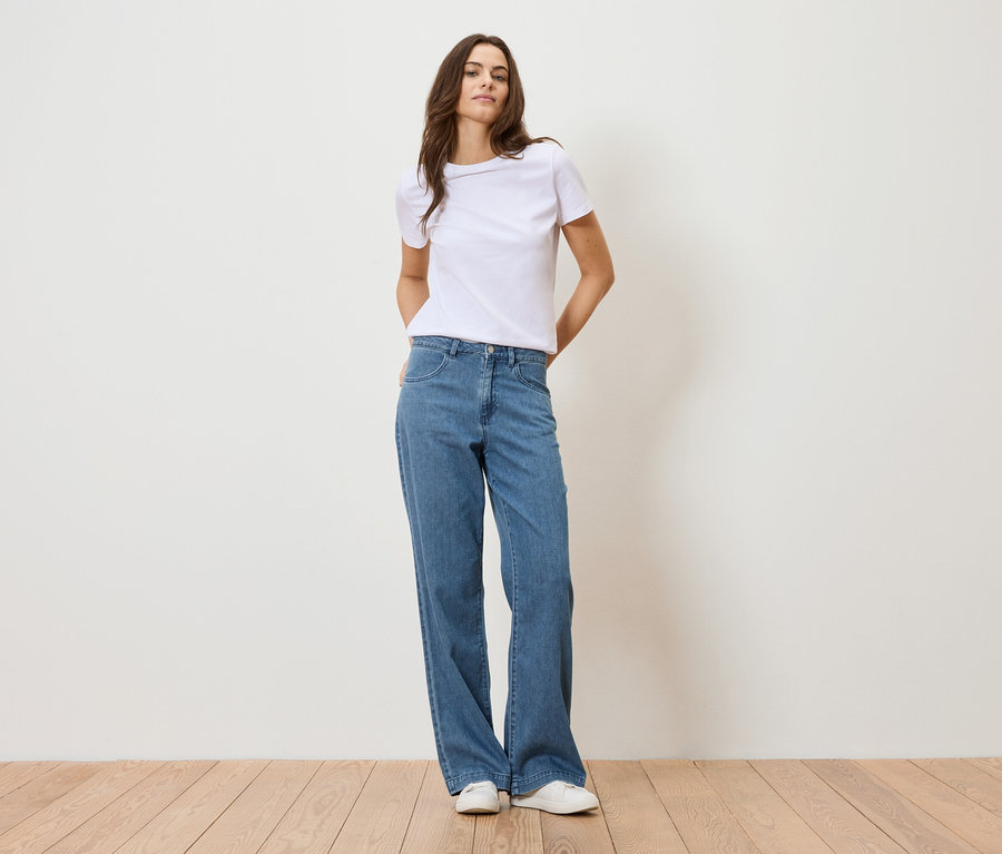 Frau steht vor einer weißen Wand und trägt ein weißes T-Shirt und blaue Jeans.