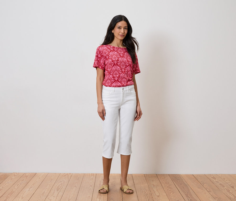 Frau steht vor einer Wand und trägt ein rotes Oberteil und weiße Hosen.