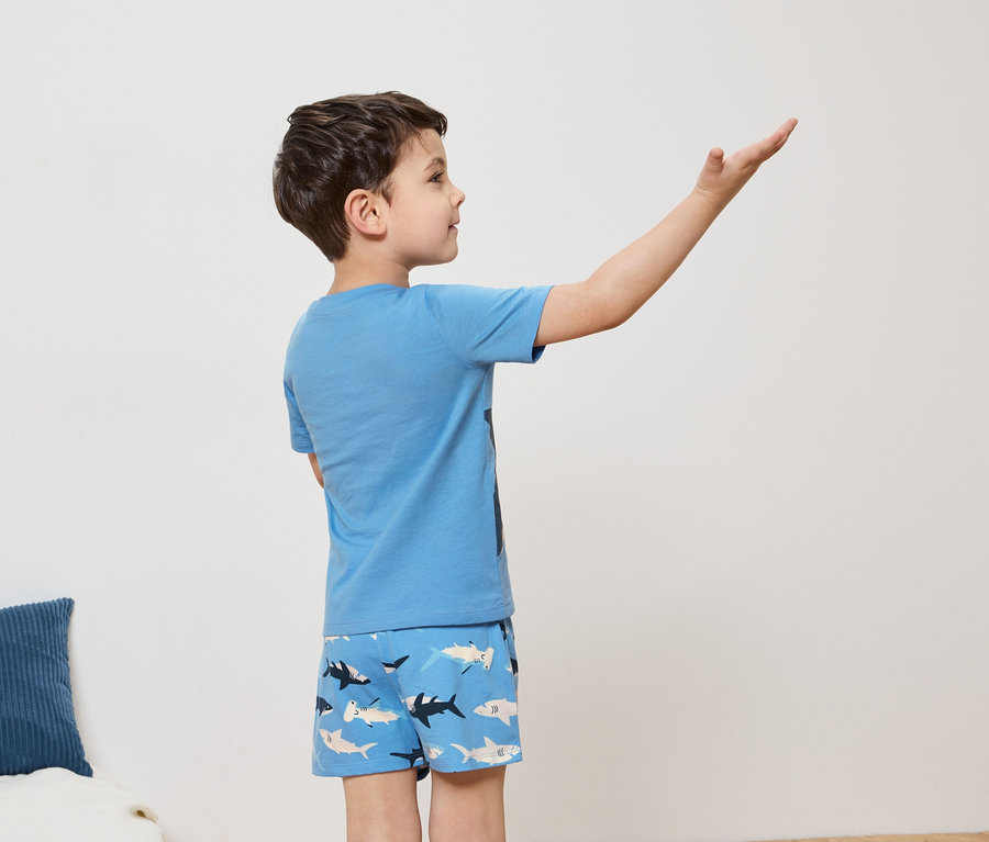 Ein Junge mit braunen Haaren, hellblauem T-Shirt und Shorts mit Hai-Muster steht seitlich mit erhobenem Arm.