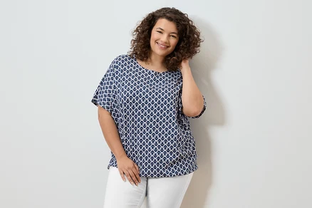 Frau mit lockigem Haar, die eine blau-weiße Bluse und weiße Hosen trägt und lächelt.