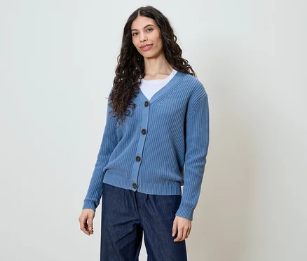Frau mit langen, dunklen, lockigen Haaren, die eine blaue Strickjacke und dunkle Jeans trägt.