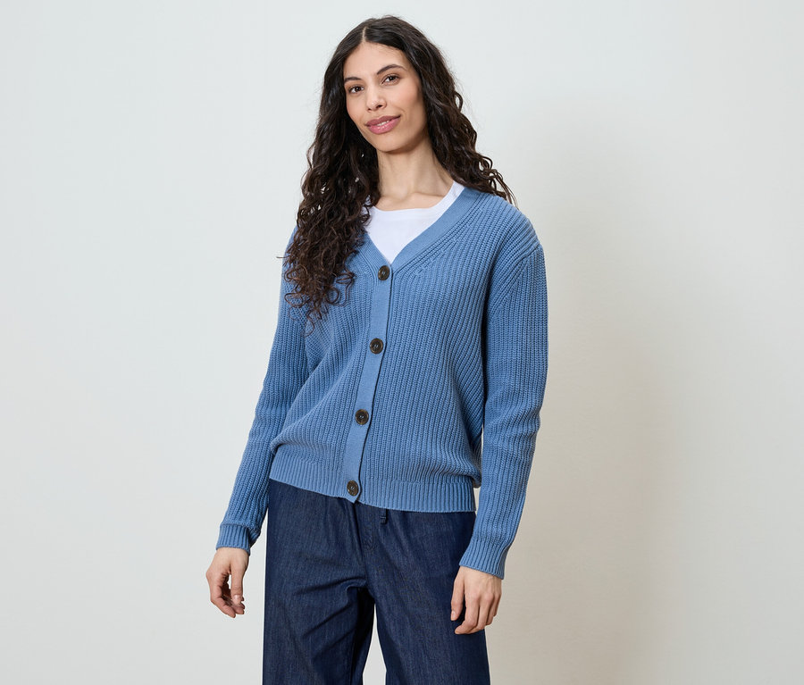 Frau mit langen, dunklen, lockigen Haaren, die eine blaue Strickjacke und dunkle Jeans trägt.