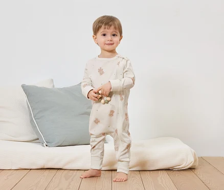 Ein Kleinkind steht barfuß vor einem Bett mit Kissen und hält eine Holzkette. Es trägt einen hellen Teddy-Overall mit Reißverschluss.
