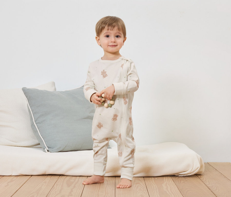 Ein Kleinkind steht barfuß vor einem Bett mit Kissen und hält eine Holzkette. Es trägt einen hellen Teddy-Overall mit Reißverschluss.