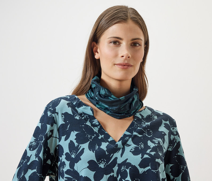 Eine Frau trägt ein blaues Oberteil mit Blumenmuster und einen passenden Schlauchschal vor einem weißen Hintergrund.