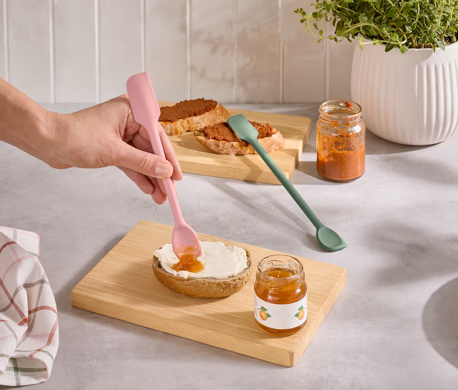 Eine Hand streicht mit einem rosa Spatel Marmelade auf ein Brötchen. Daneben stehen ein grüner Spatel und zwei Marmeladengläser.