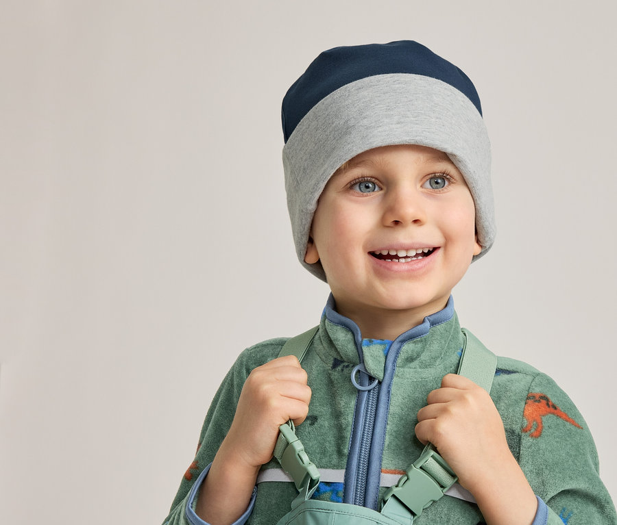 Lächelnder Junge mit blau-grauer Mütze und grünem Dino-Overall hält Hosenträger.