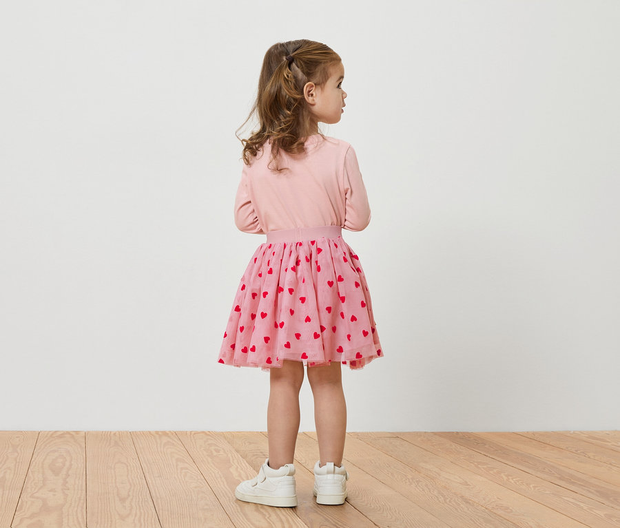 Mädchen von hinten in rosa Oberteil und Rock mit roten Herzen, dazu weiße Schuhe.