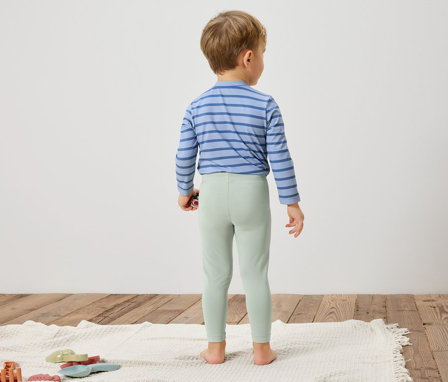 Ein Kind steht mit dem Rücken zur Kamera, trägt ein blau gestreiftes Hemd und hellgrüne Leggings.