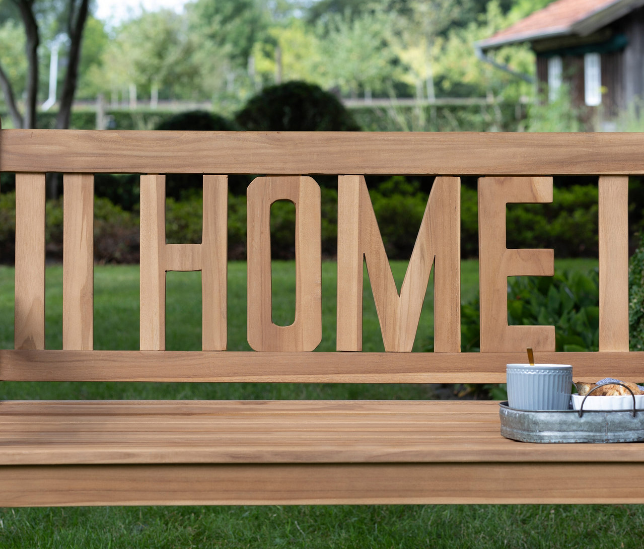 Eine deVries Gartenbank »Home« steht im Garten, darauf eine Tasse und ein Teller auf einem Tablett.