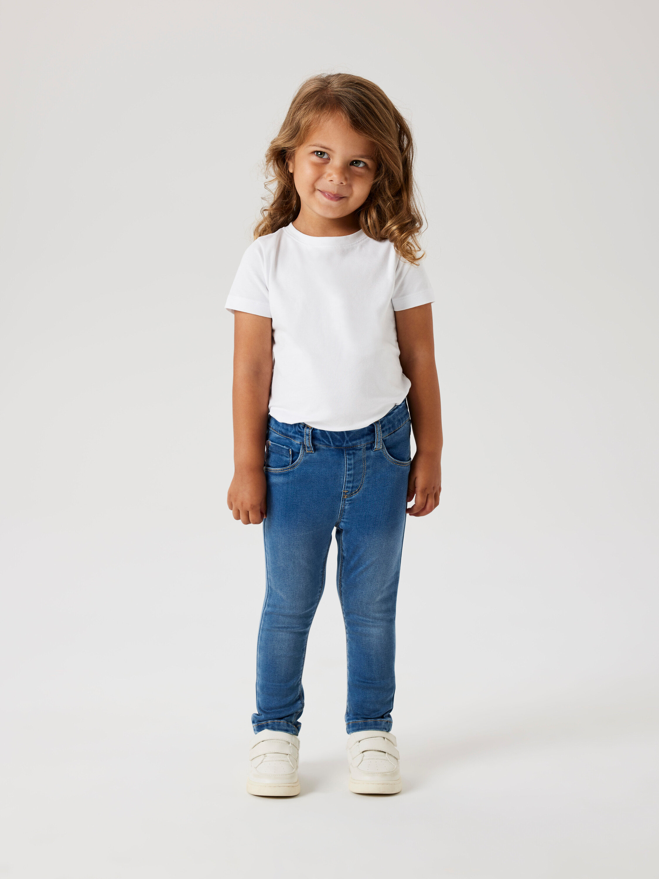 Mädchen mit lockigem Haar trägt ein weißes T-Shirt, blaue Jeans und weiße Schuhe.