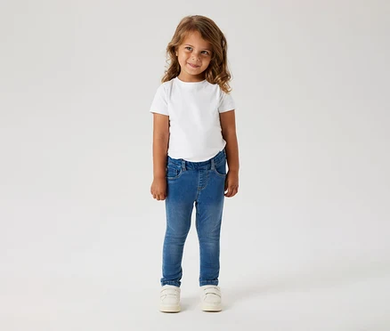 Mädchen mit lockigem Haar trägt ein weißes T-Shirt, blaue Jeans und weiße Schuhe.