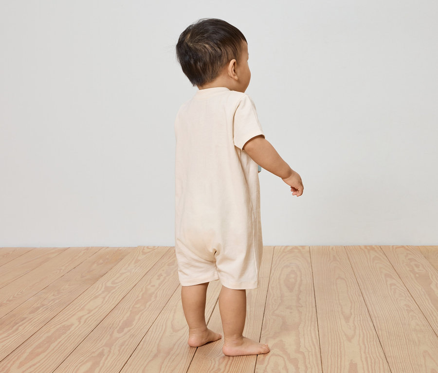 Ein Kind steht in einem hellbeigen Kurzarm-Overall auf einem Holzboden, mit dem Rücken zur Kamera.