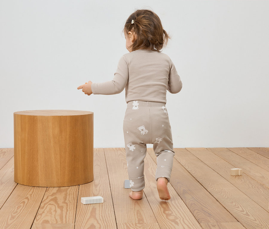 Kleines Kind in beiger Kleidung mit Bärenmuster zeigt auf einen Holzhocker und Spielzeug auf einem Holzboden.