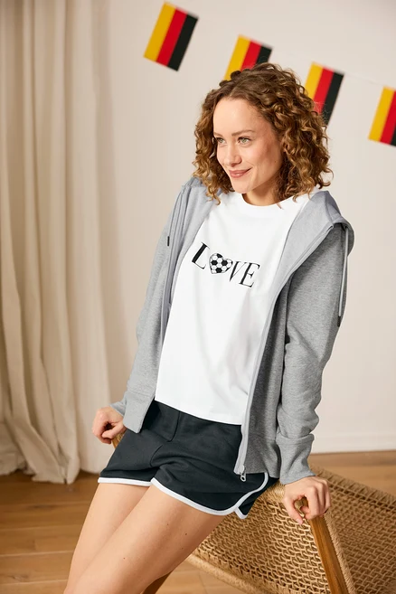 Frau mit Sweatshorts, Sportshirt mit Fußballmotiv und grauer Sweatjacke sitzt vor Deutschland-Girlande.