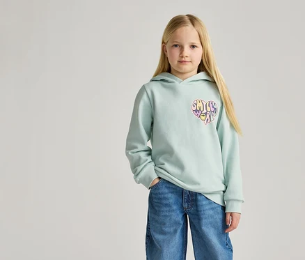 Mädchen mit blonden Haaren trägt hellblauen Kapuzenpullover mit Herz-Smiley-Aufdruck und Jeans.